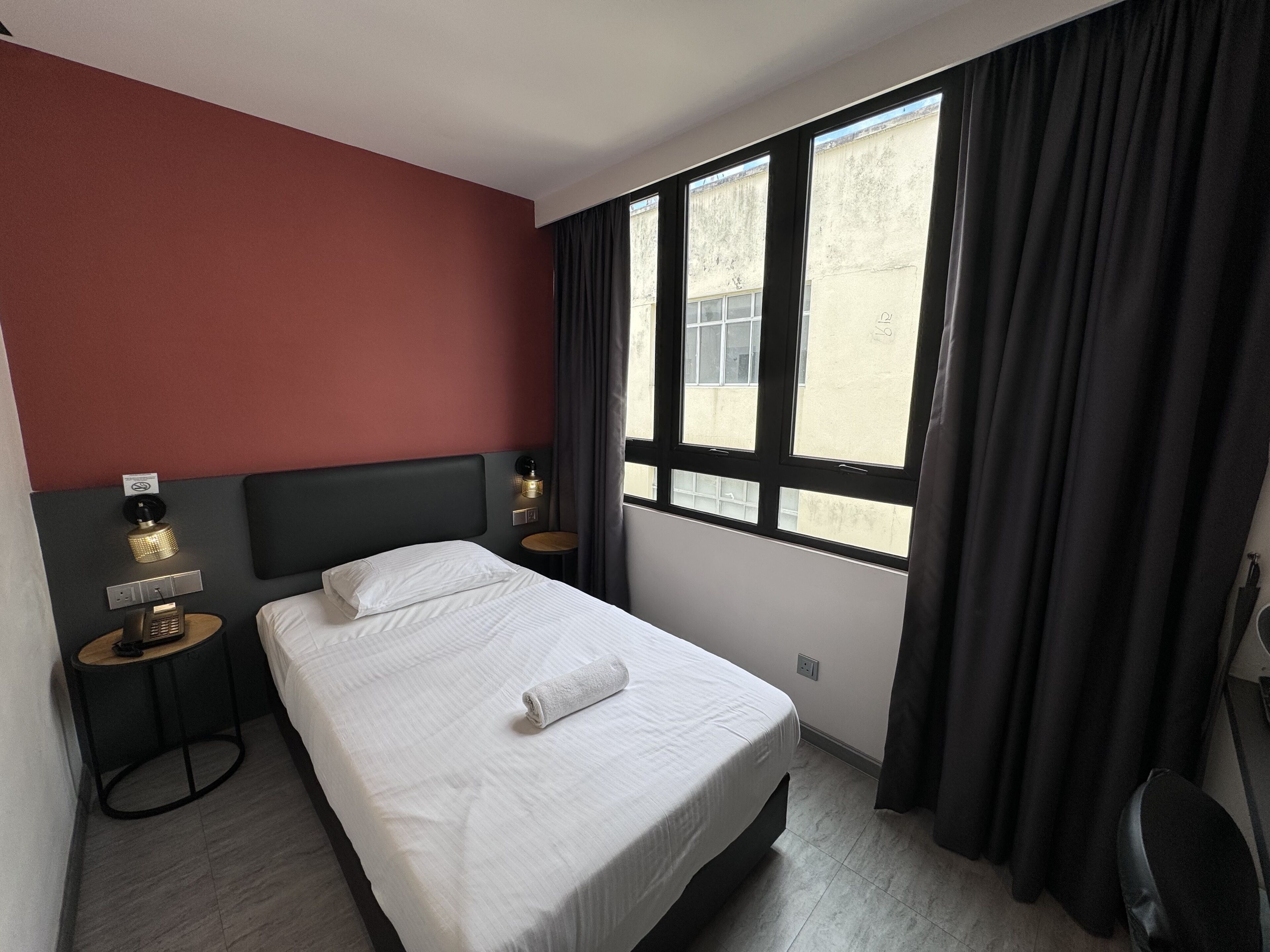 A hotel room with a bed, two bedside tables, a wall-mounted lamp, a window with curtains, and a view of the outside.
