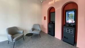 Un quiosco de auto-check-in, dos sillones y una mesita en una habitación con pared rosa y una lámpara en el techo.