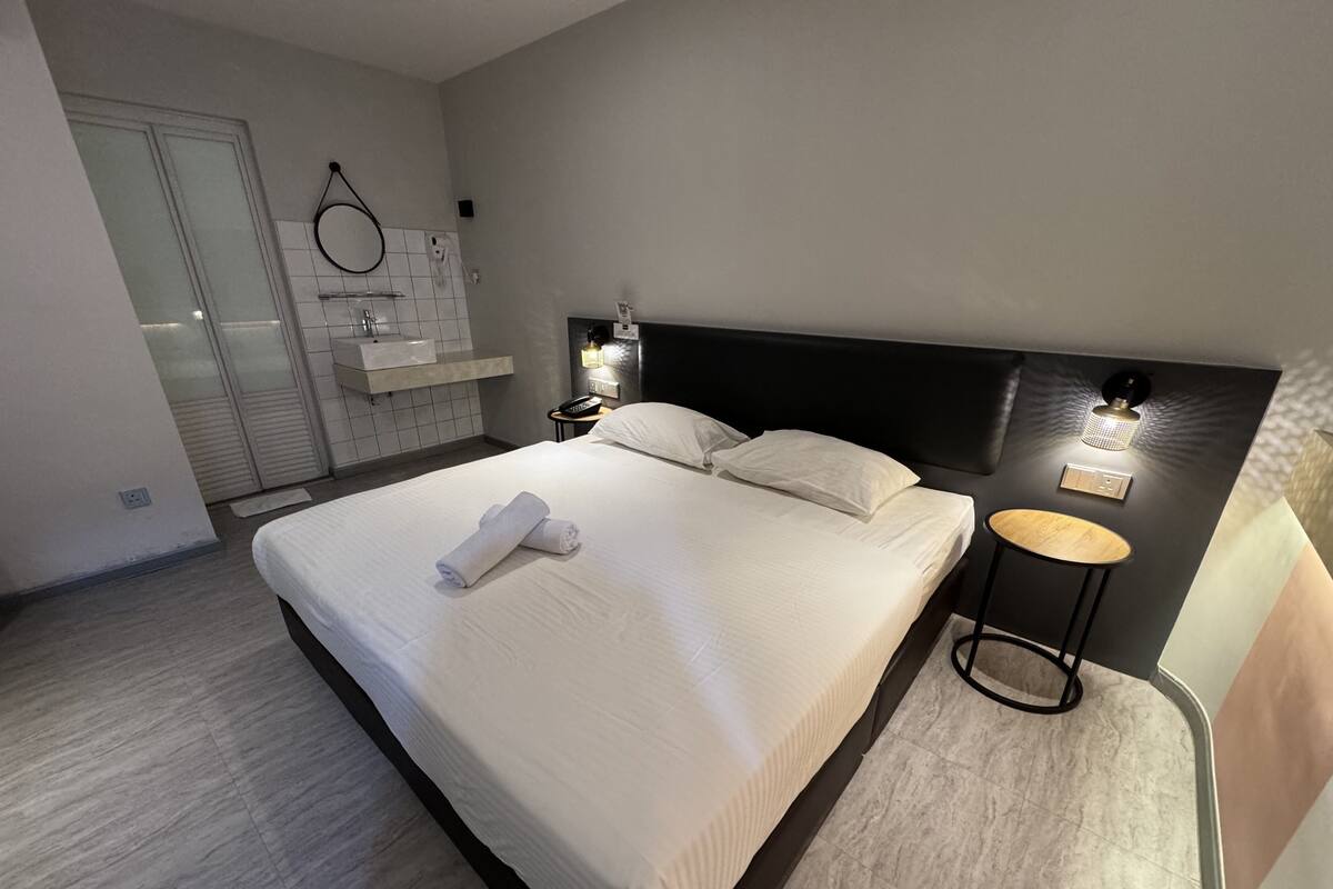 Una habitación de hotel moderna con una cama grande, una mesa redonda, una mesita de noche y un baño con espejo y lavamanos.