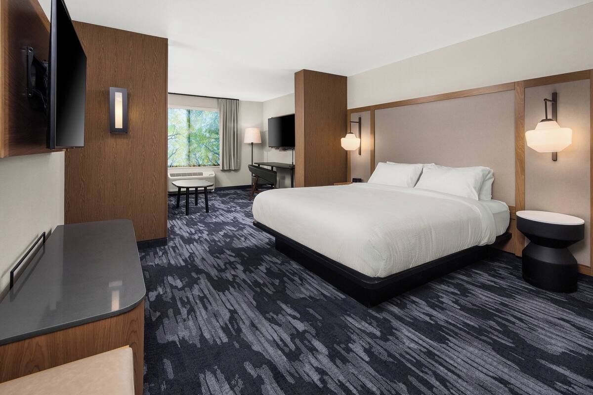 A hotel room with a large bed, a desk, a chair, a TV, and a window with a view of trees.