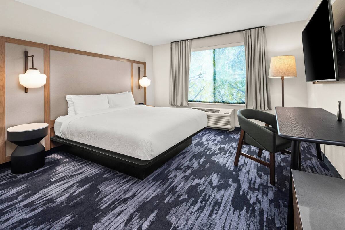 A hotel room with a large bed, a desk, a chair, a TV, and a window with curtains.