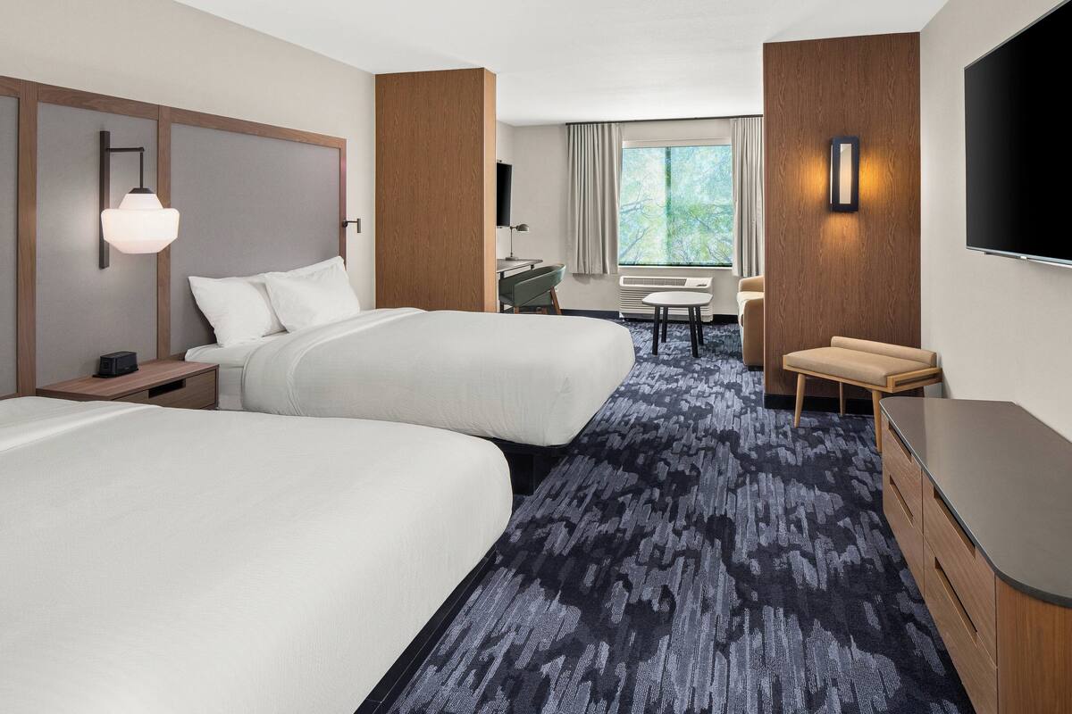 A hotel room with two beds, a desk, a chair, a TV, and a large window.