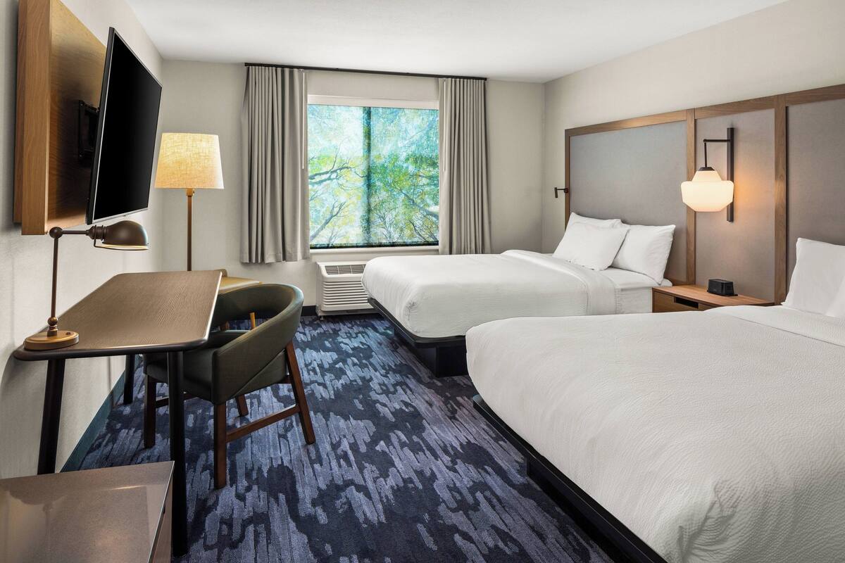 A hotel room with two beds, a desk with a chair, a flat-screen TV, a window with curtains, and a wall-mounted lamp.