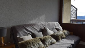 Un sofá gris con varios cojines, una lámpara sobre una mesita de madera y vista a un edificio a través de una ventana.