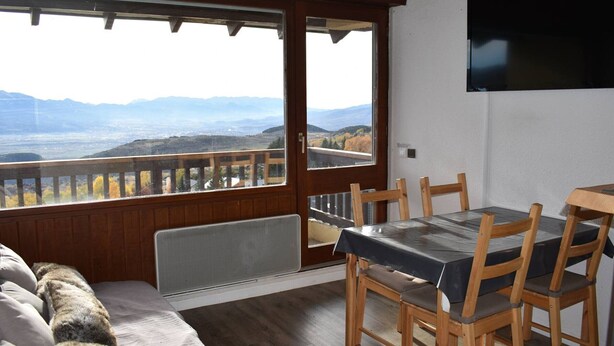Una habitación con piso de madera, un sofá con una manta de piel, una mesa de comedor con sillas, un televisor montado en la pared y un amplio ventanal con vistas a montañas.