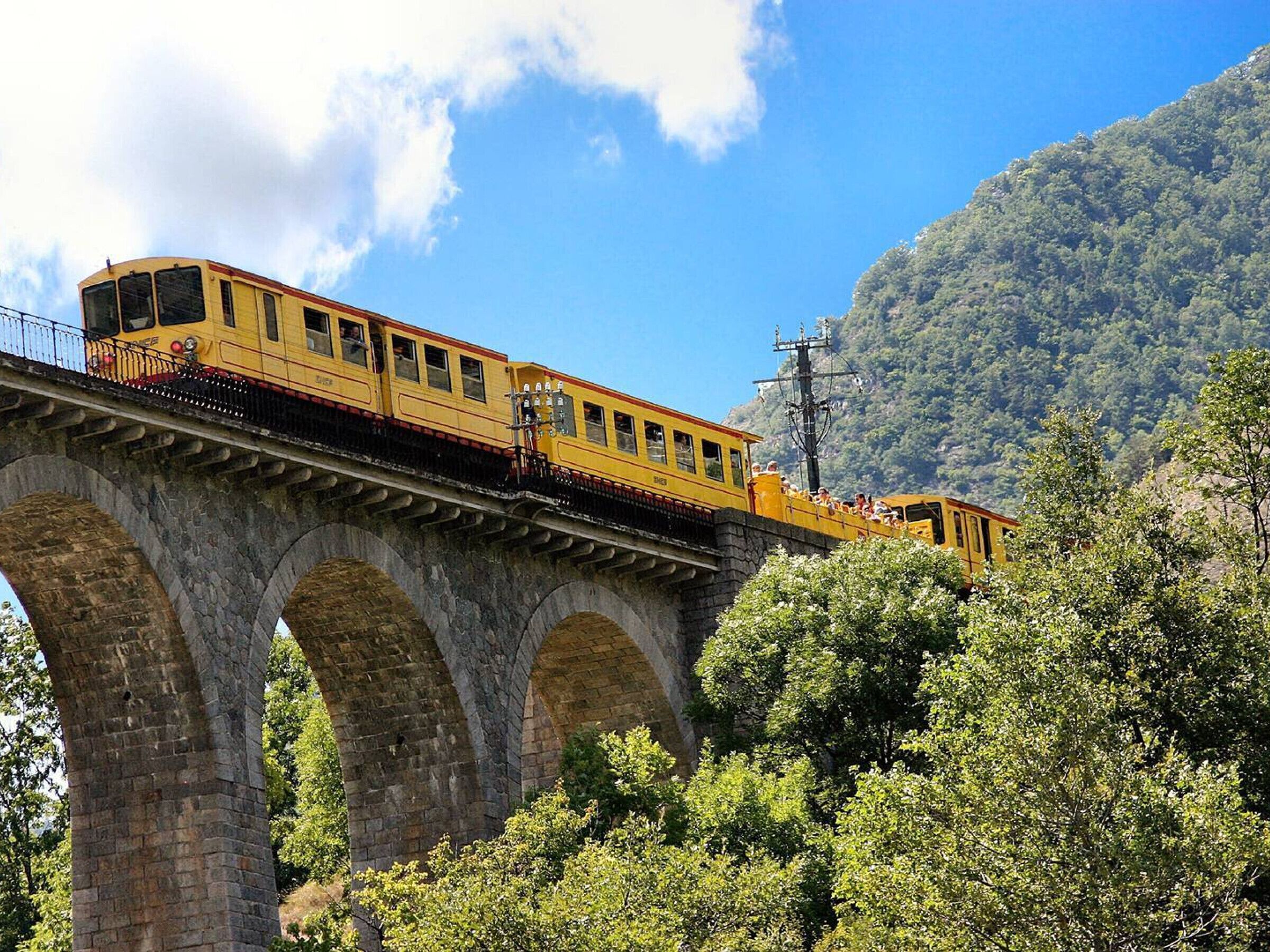 A yellow train on a stone bridge with arched supports, surrounded by greenery and a mountainous backdrop.