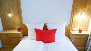 Una cama con ropa de cama blanca y un almohárraga rojo, flanqueada por mesitas de noche de madera con pequeñas macetas.