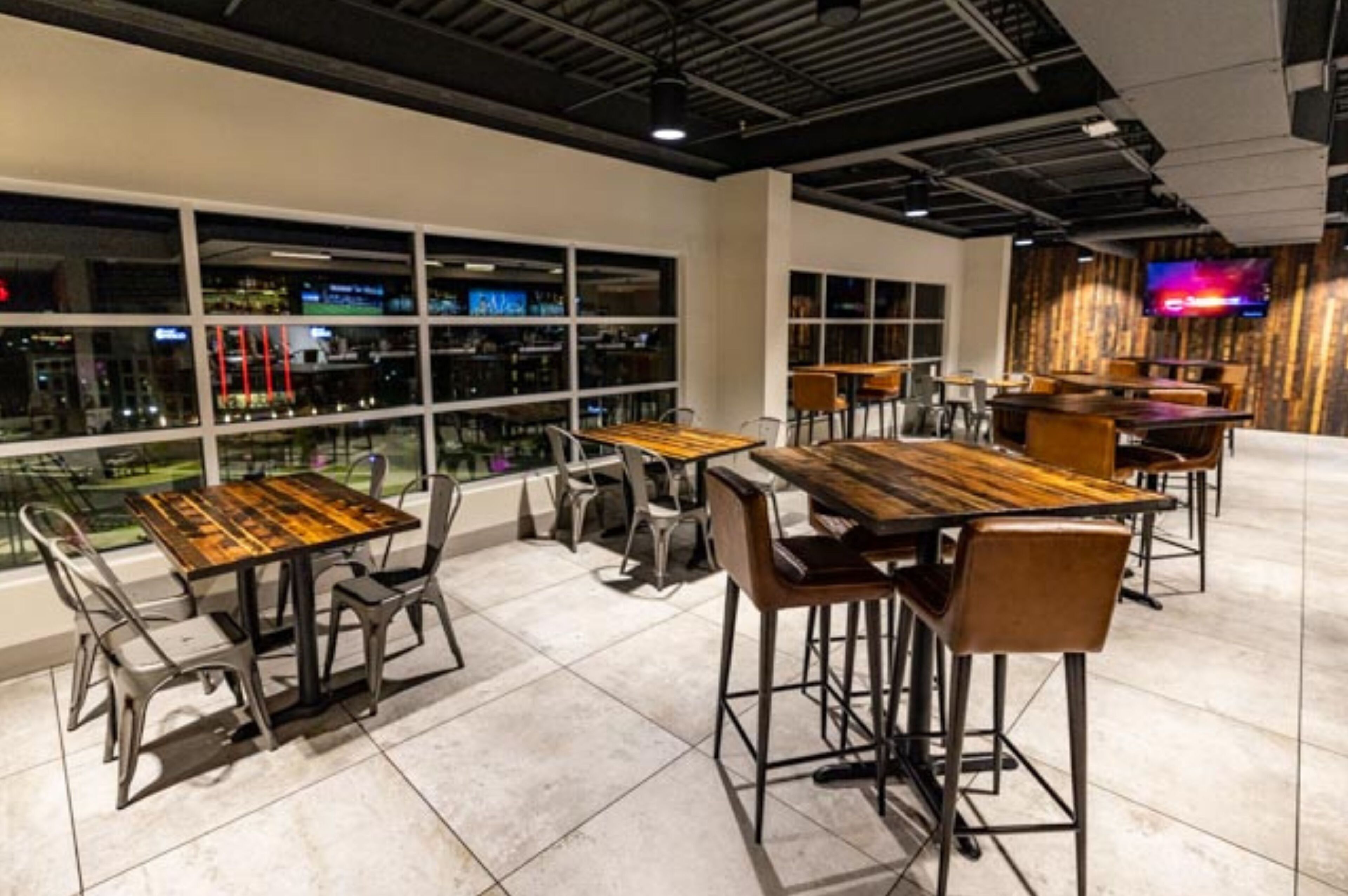 A modern restaurant with high tables and chairs, large windows, and a TV on the wall.