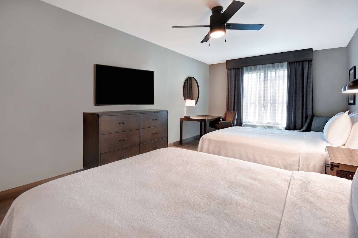 A hotel room with two beds, a flat-screen TV, a ceiling fan, a dresser, a desk, and a window with curtains.
