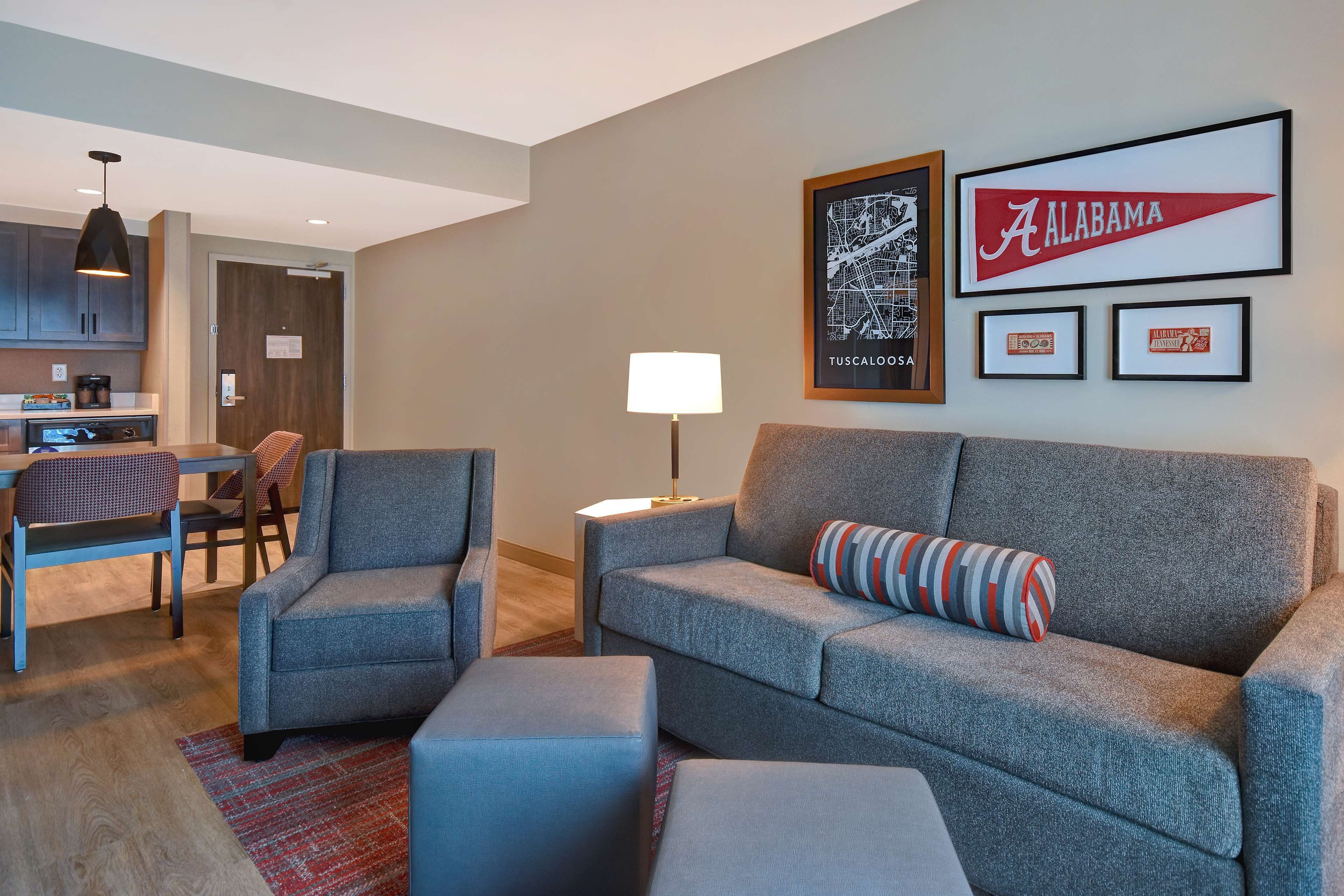 A modern living room with a grey sofa, armchairs, and a coffee table. There is a kitchen area with cabinets and a dining table. The walls feature framed artwork, including a map and a sign reading "Alabama."