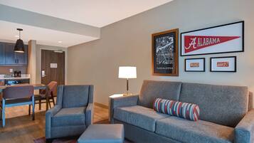 A modern living room with a grey sofa, armchairs, and a coffee table. There is a kitchen area with cabinets and a dining table. The walls feature framed artwork, including a map and a sign reading "Alabama."