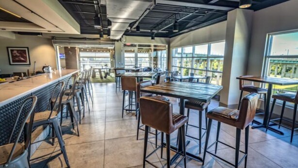 A modern bar area with high tables and stools, large windows, and a view of the cityscape. - Homewood Suites by Hilton Tuscaloosa Downtown (Tuscaloosa)