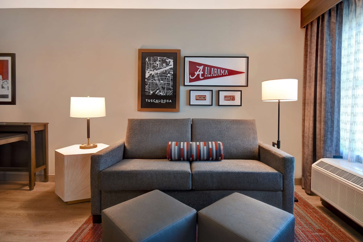 A modern hotel room with a grey sofa, two side tables with lamps, framed wall art, and a window with curtains.