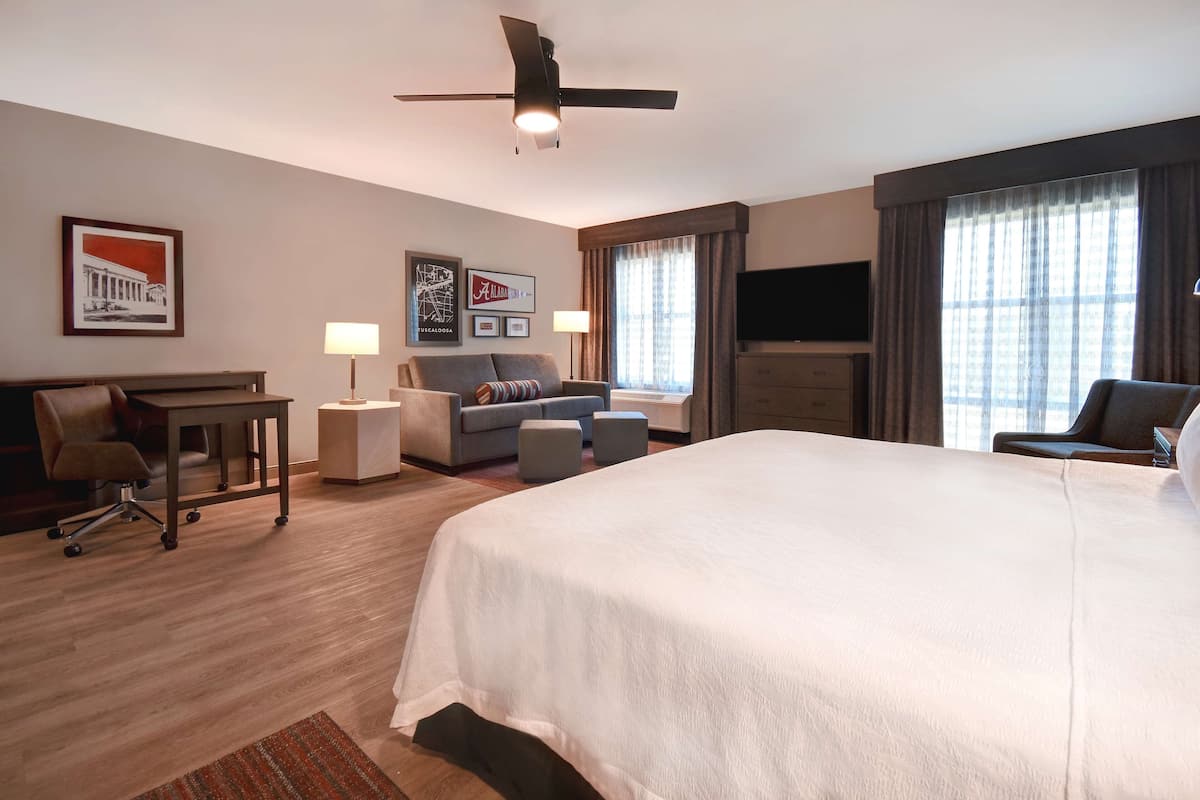 A hotel room with a large bed, a desk with a chair, a sofa, two side tables, a ceiling fan, a flat-screen TV, and framed pictures on the wall.
