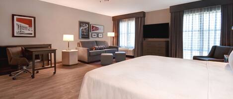 A hotel room with a large bed, a desk with a chair, a sofa, two side tables, a ceiling fan, a flat-screen TV, and framed pictures on the wall.