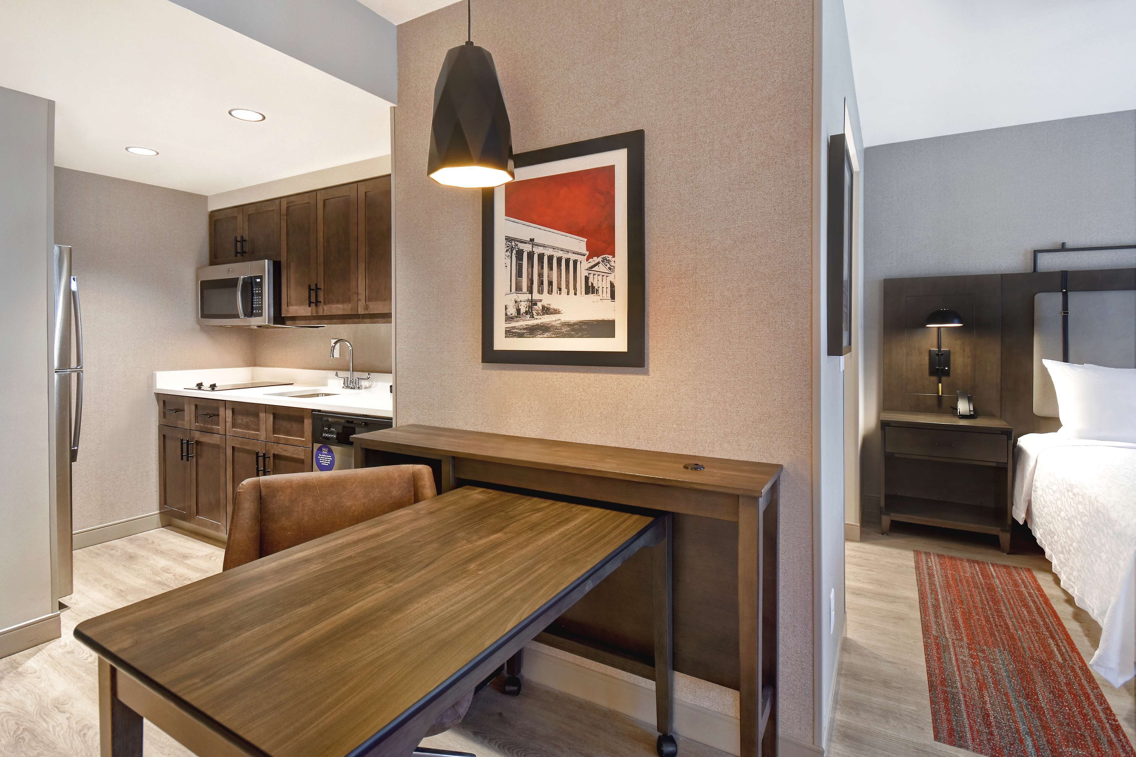 Una habitación de hotel moderna con una mesa de comedor de madera, una zona de cocina con microondas y una cama con mesita de noche.