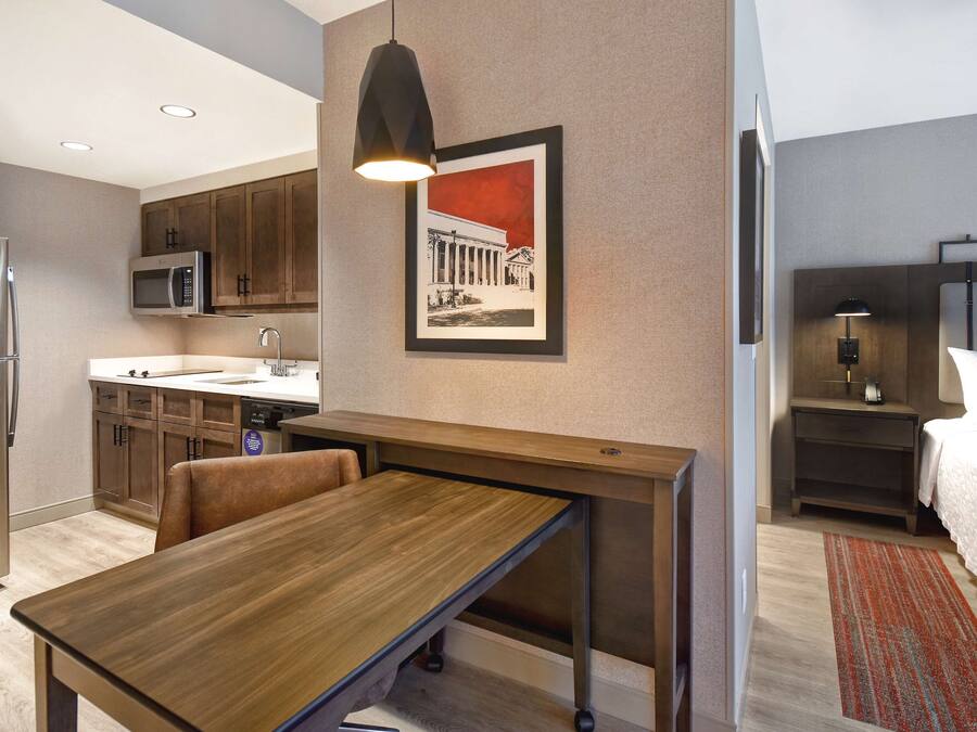 Una habitación de hotel moderna con una mesa de comedor de madera, una zona de cocina con microondas y una cama con mesita de noche.