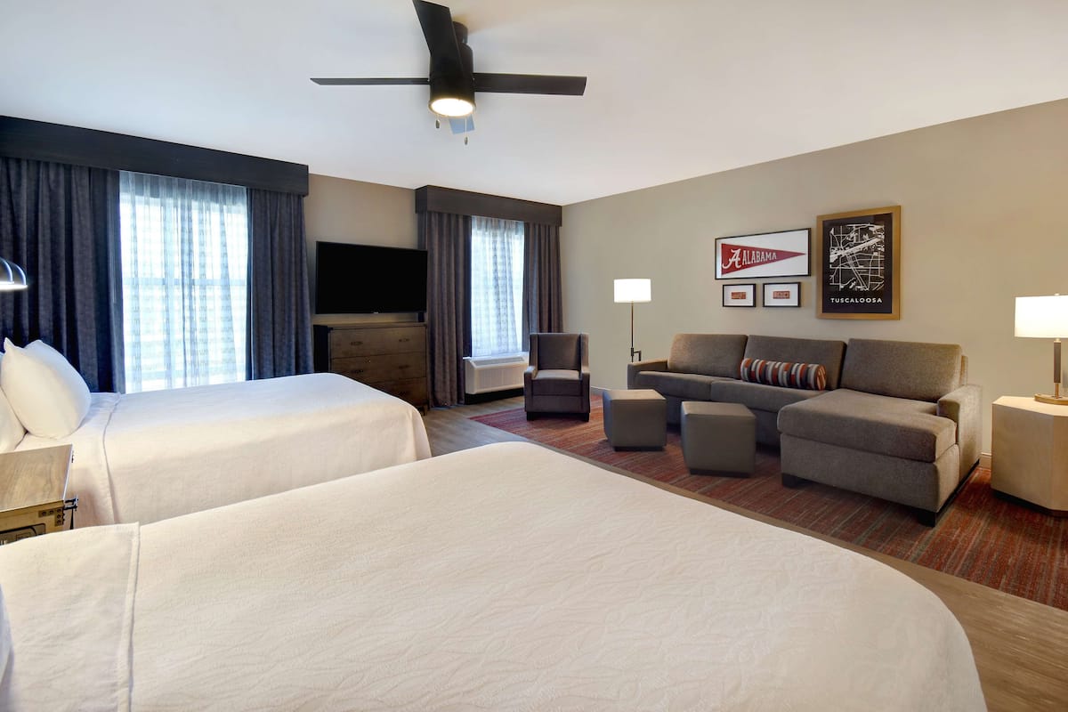 A hotel room with two beds, a sofa, a chair, a TV, and a ceiling fan.