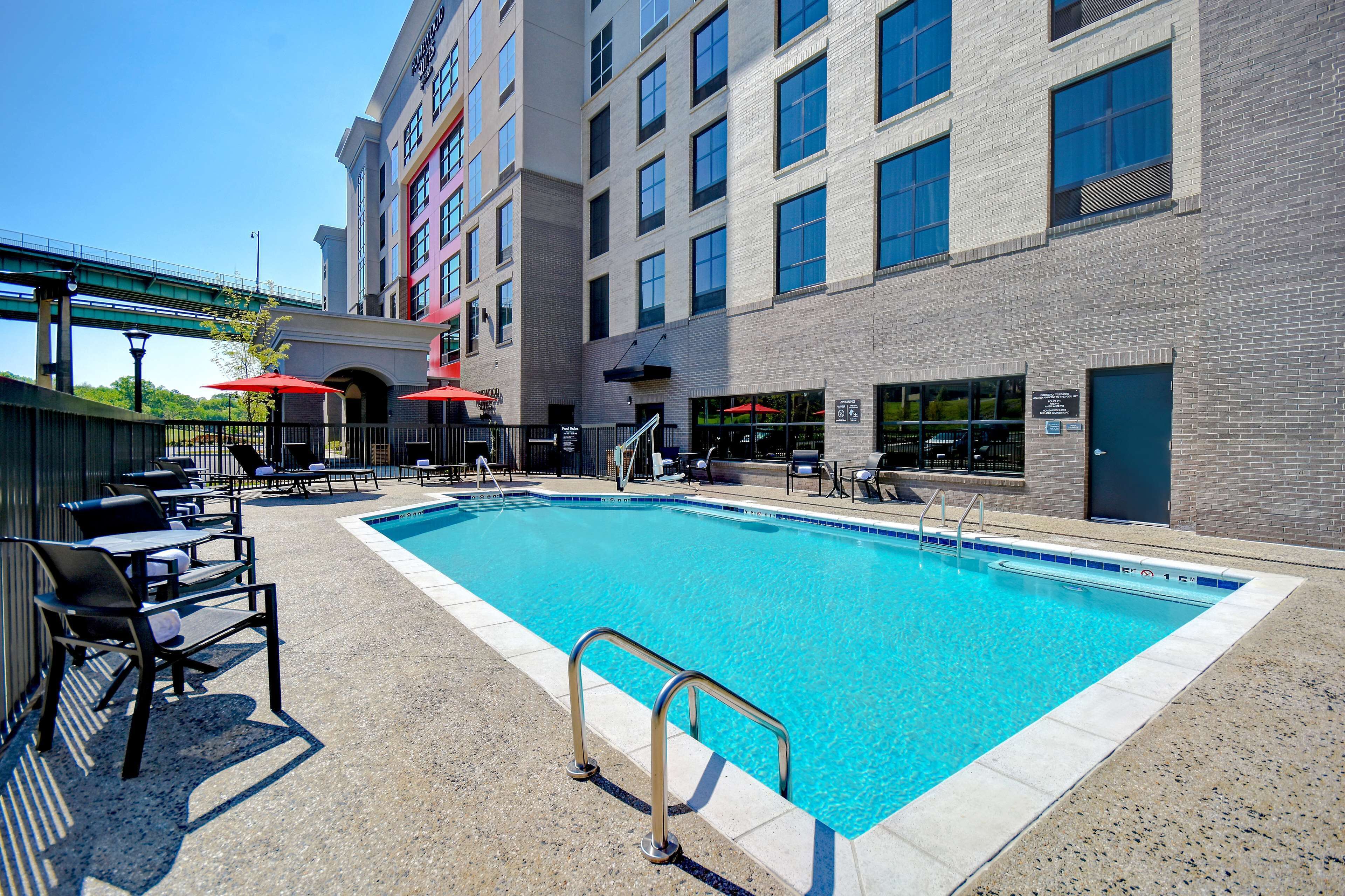 A swimming pool with lounge chairs and umbrellas, surrounded by a brick building with multiple windows and a covered walkway.