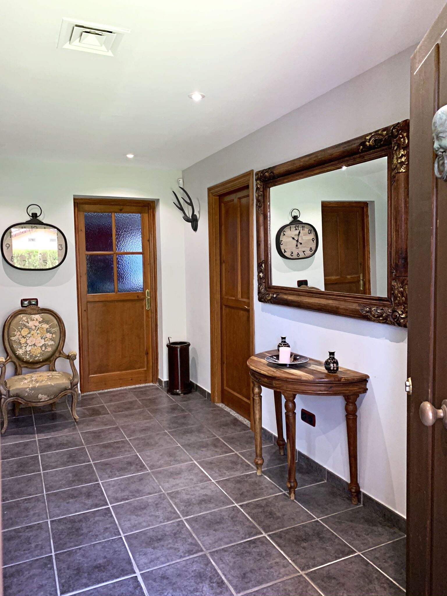 Een gang met een houten deur, een spiegel, een klok, een stoel en een tafeltje met decoratieve objecten.