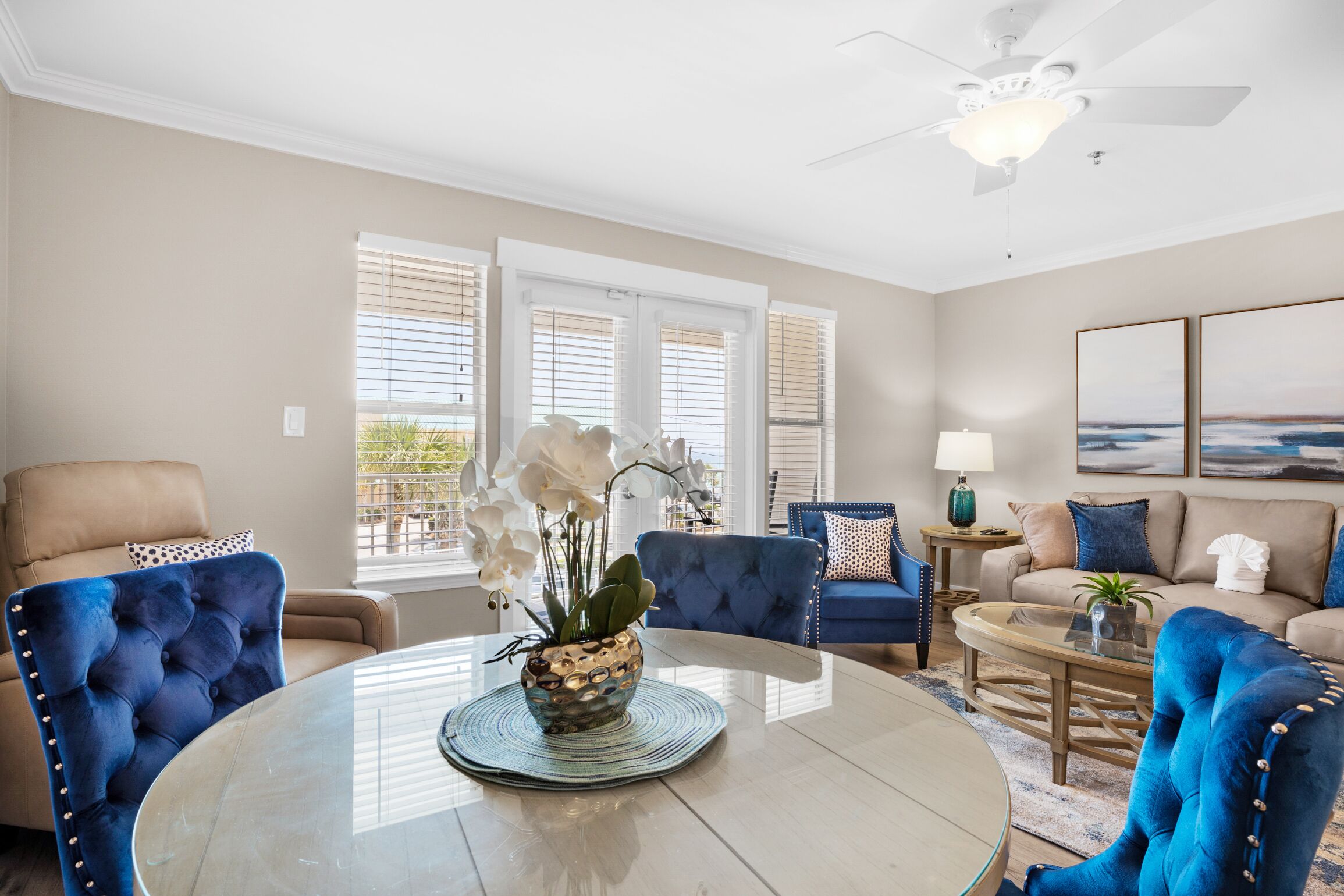 A modern living room with a round glass coffee table, blue upholstered chairs, a beige sofa, and a ceiling fan.