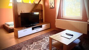 Un soggiorno con televisore a schermo piatto, un tavolino, un divano, una finestra con tende e un termosifone.