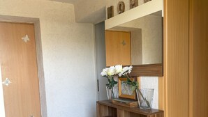 Una stanza con una porta in legno, un parete bianca, uno specchio, un comò in legno con cassetti e un piccolo vaso con fiori bianchi.