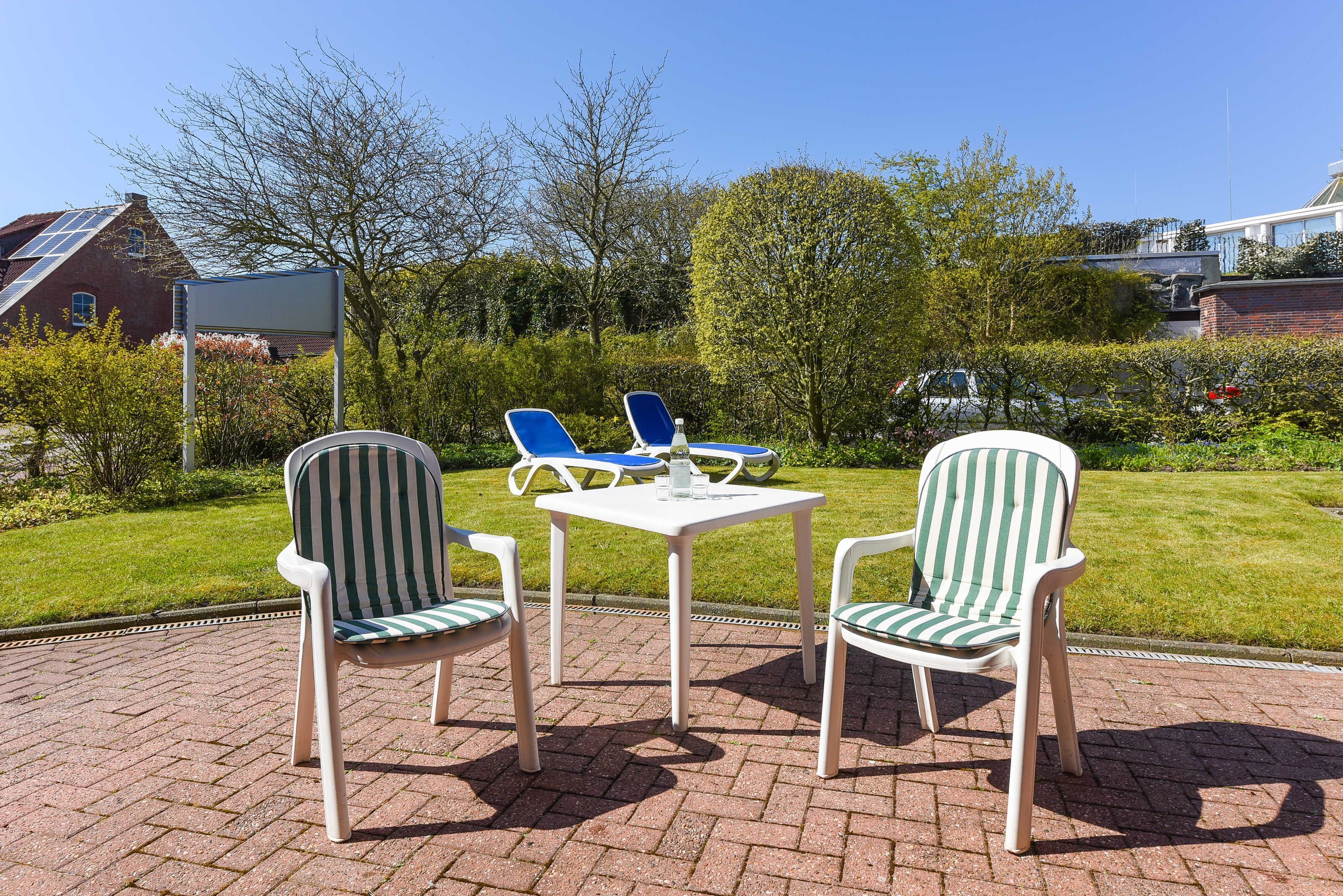 Une terrasse avec deux chaises à rayures et une table, entourée d’un chemin en briques et de verdure.
