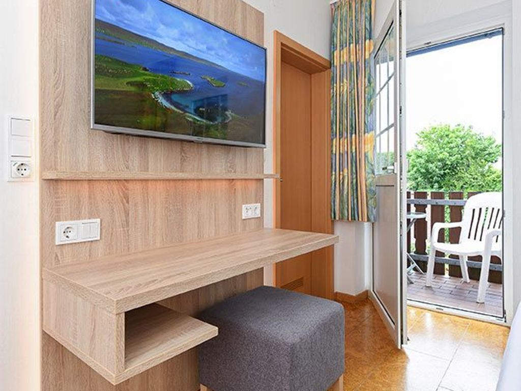 Un salon moderne avec un téléviseur à écran plat fixé à un mur en bois, un petit banc gris et une porte-fenêtre coulissante donnant sur un espace extérieur où se trouve une chaise blanche.