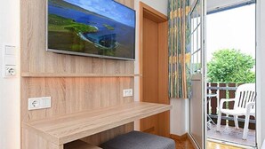 Ein modernes Wohnzimmer mit einem Flachbildfernseher an einer Holzwand, einer kleinen grauen Bank und einer Glastür, die zu einem Außenbereich mit einem weißen Stuhl führt.
