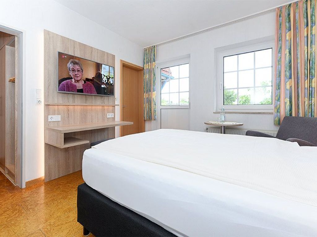 Habitación de hotel con una cama grande, un televisor montado en un panel de madera, una mesita y una ventana con cortinas.
