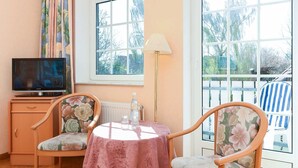 Een gezellige kamer met een televisie, een tafel met een roze tafelkleed, twee bloemenstoelen, een houten kast en een raam met uitzicht op bomen.