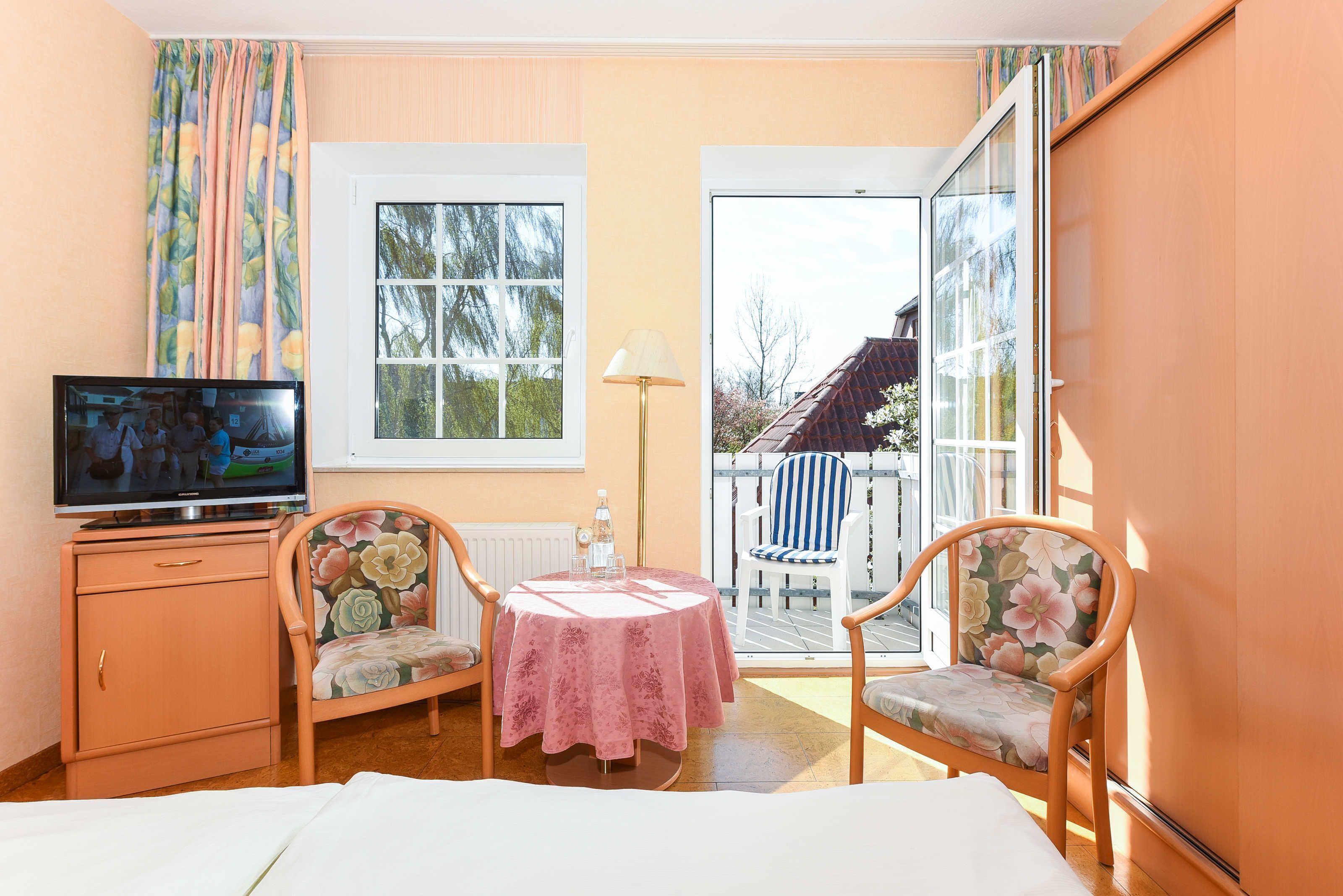 Ein Zimmer mit einem Bett, einem Fernseher, einem Tisch mit rosa Tischdecke, zwei Stühlen mit floralem Bezug, einer Lampe und Blick auf einen Garten durch eine Schiebetür.