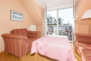 A room with a table covered by a pink tablecloth, two striped armchairs, a radiator, and a large window with a view of the outdoors.