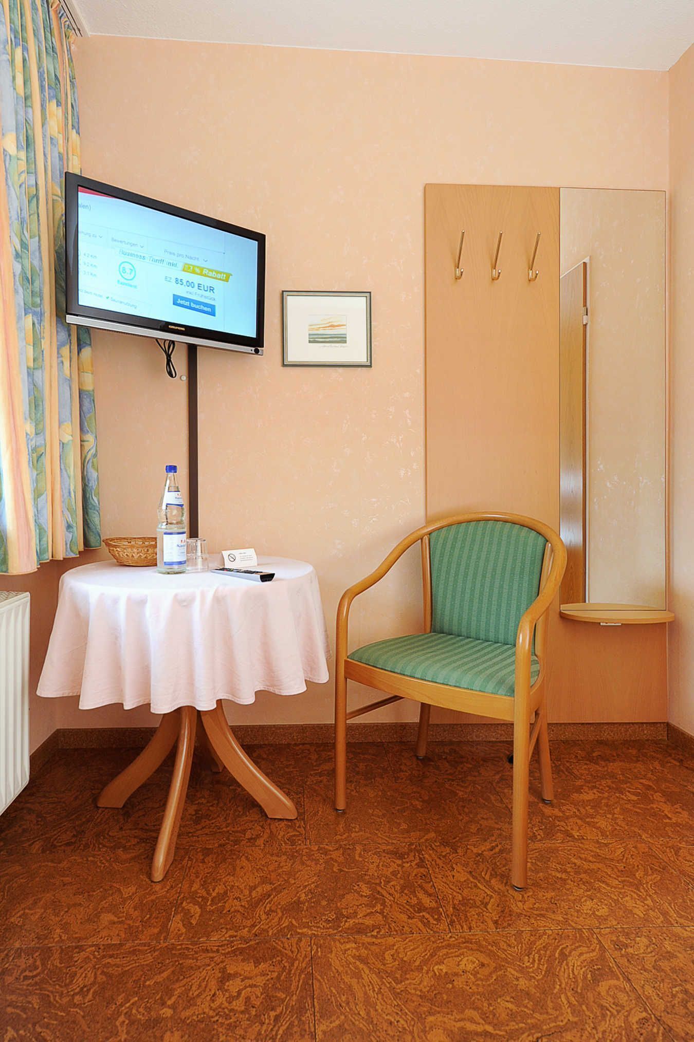 A hotel room with a television on a stand, a table with a bottle and glass, a chair, and a wardrobe with hooks.