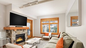 A modern living room with a stone fireplace, a flat-screen TV mounted above it, a sofa with orange cushions, and a window with a view of trees. - Incredible Snowmass Condo by Gondola! Walk to Dining & Shops. A/C, Hot Tubs, Balcony, W/D, Parking! (Snowmass Village)