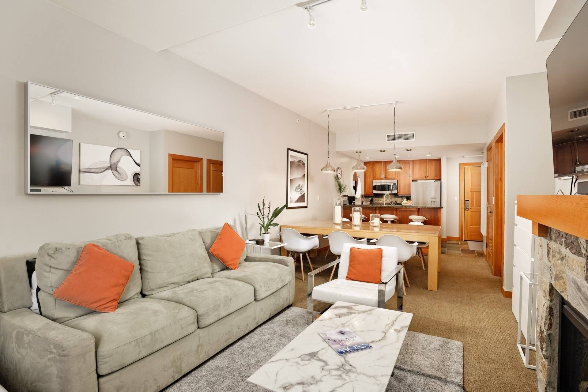 Un salon moderne avec un canapé gris, des coussins orange, une table basse en marbre et un coin repas comprenant des chaises blanches et une table en bois.