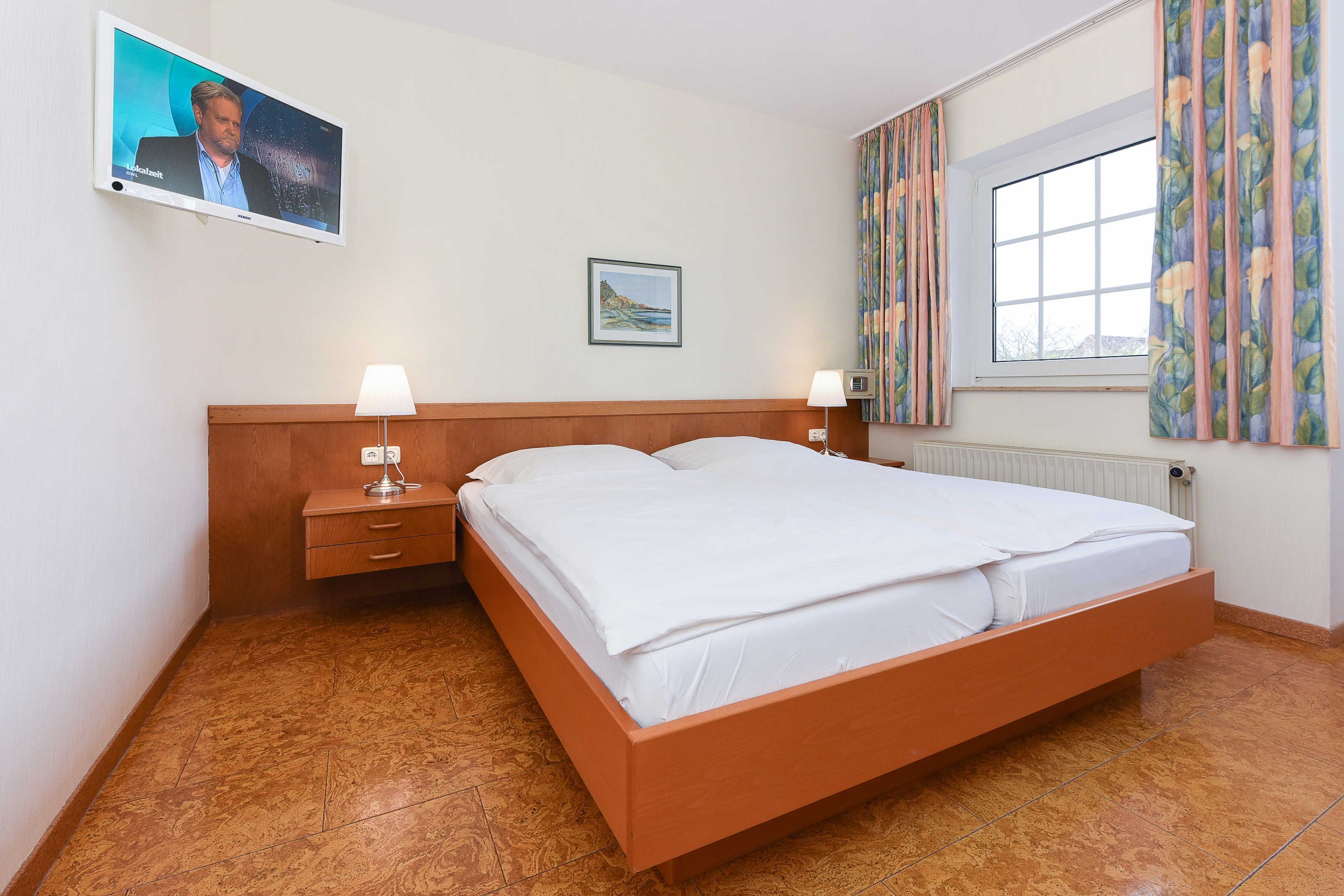 Une chambre d’hôtel avec un lit en bois, deux tables de chevet avec des lampes, un téléviseur fixé au mur, une fenêtre avec des rideaux et un tableau encadré accroché au mur.