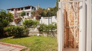Una ducha al aire libre con paredes de piedra, un área con césped y un edificio con varios balcones bajo un cielo azul despejado.