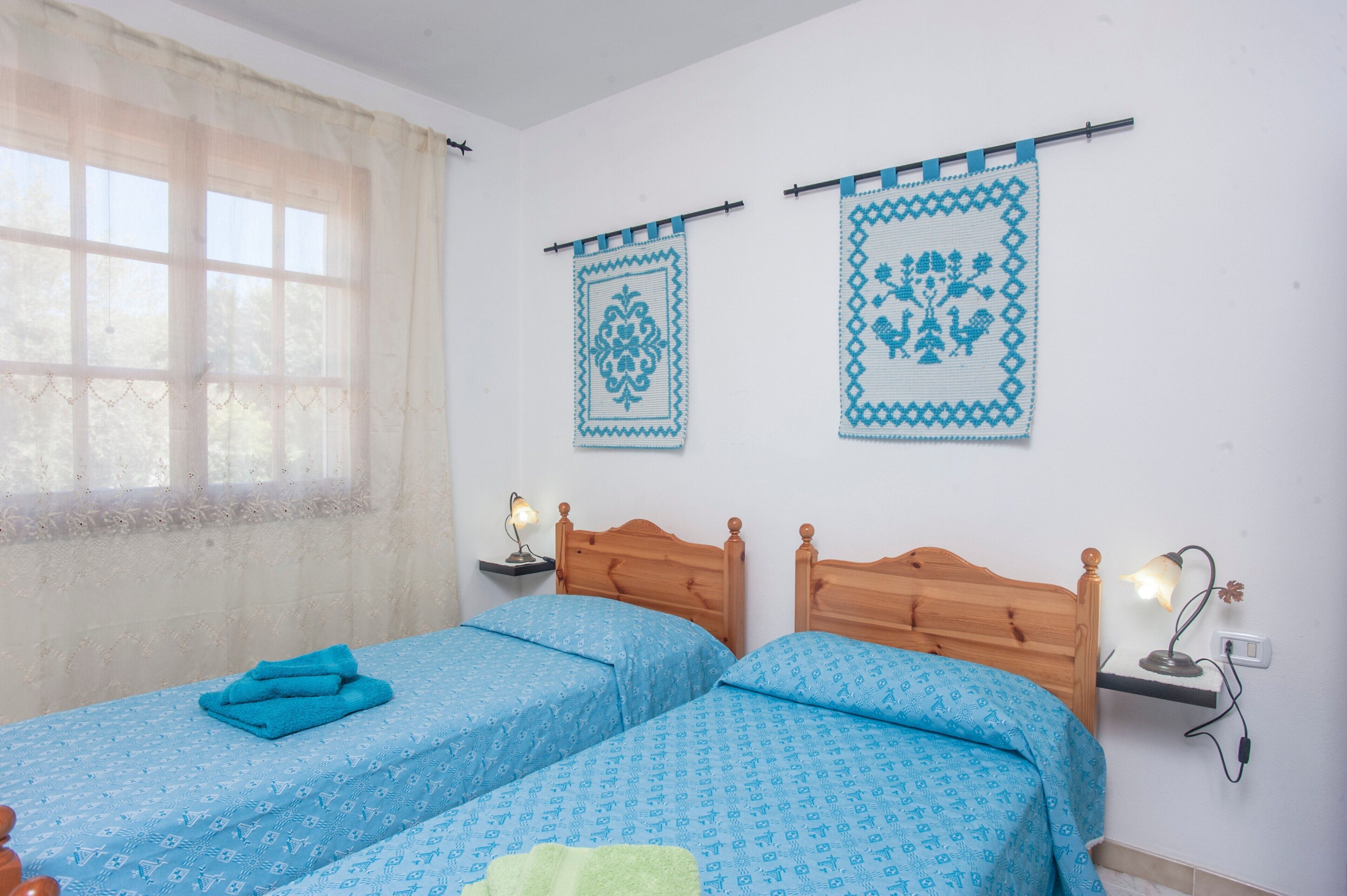 A room with two beds, a window with sheer curtains, wall-mounted lamps, and two wall hangings.