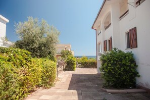A narrow paved pathway leading to a white building with red shutters, surrounded by greenery and a clear sky.