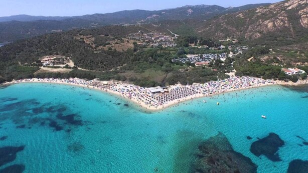 Vista aérea de una zona costera con playa, aguas turquesas cristalinas y montañas al fondo.