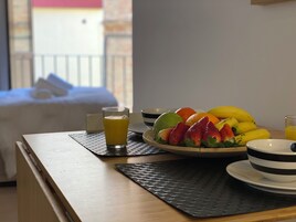 Una mesa de desayuno con un frutero, un vaso de zumo de naranja y una cama al fondo.