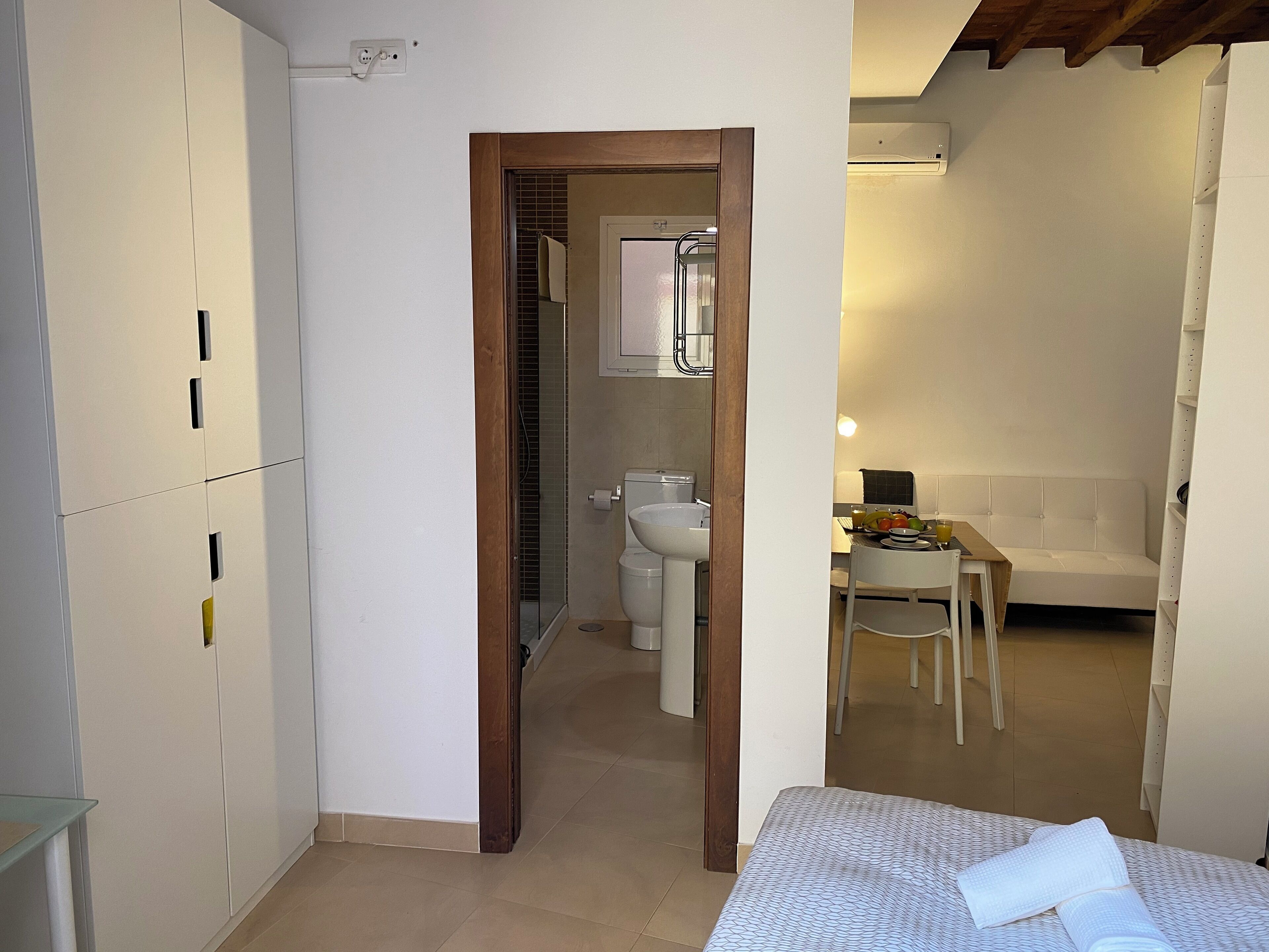 Habitación pequeña y funcional con cama, una mesita y una silla, y baño a la vista a través de una puerta abierta.
