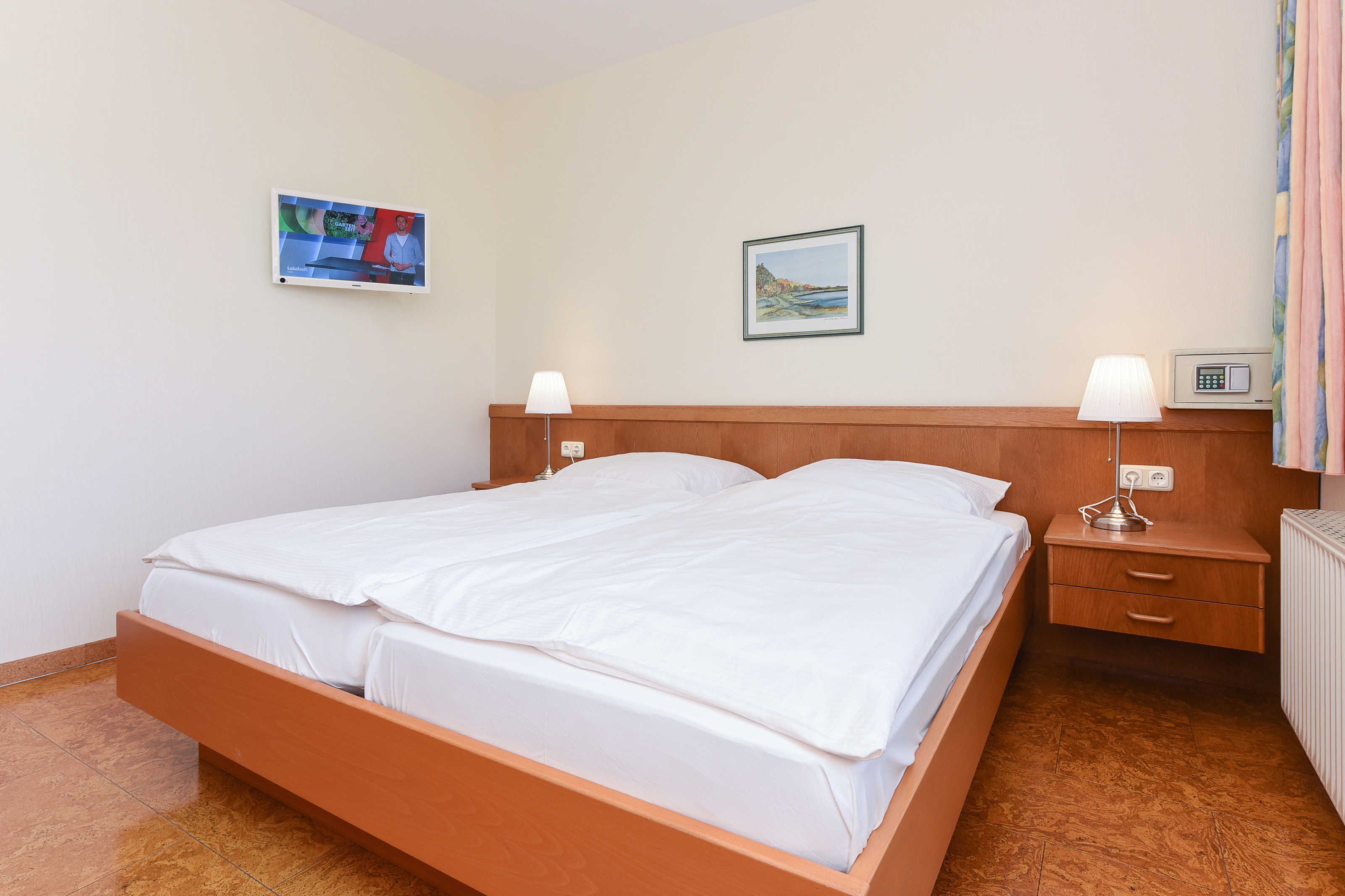 A hotel room with a wooden bed, two bedside tables, a wall-mounted TV, a framed picture, and a radiator.