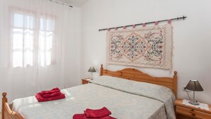 A hotel room with a bed, a nightstand, a lamp, a window with curtains, and a wall tapestry.