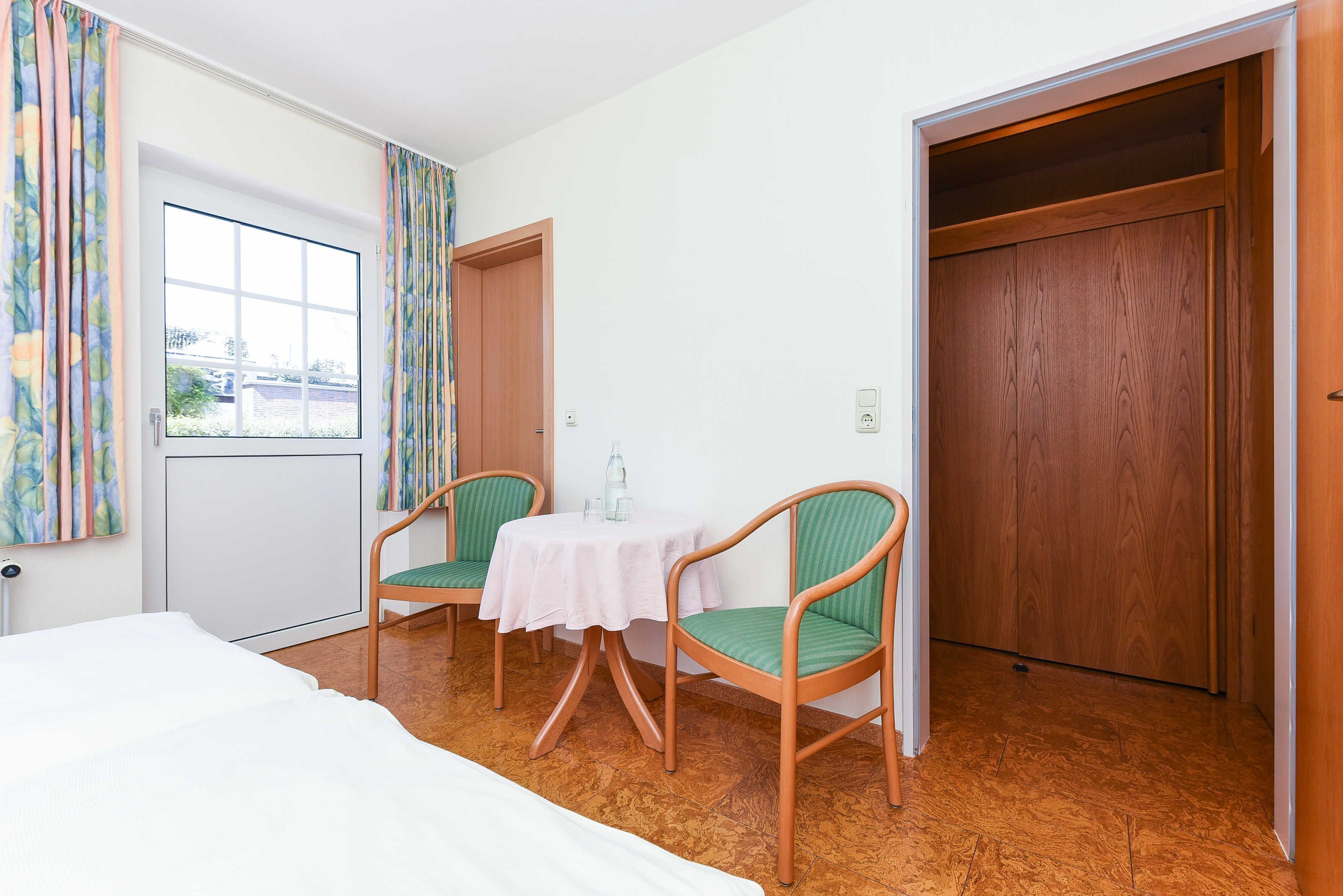 A hotel room with a bed, two chairs, a small table, a window with curtains, and a wooden door.