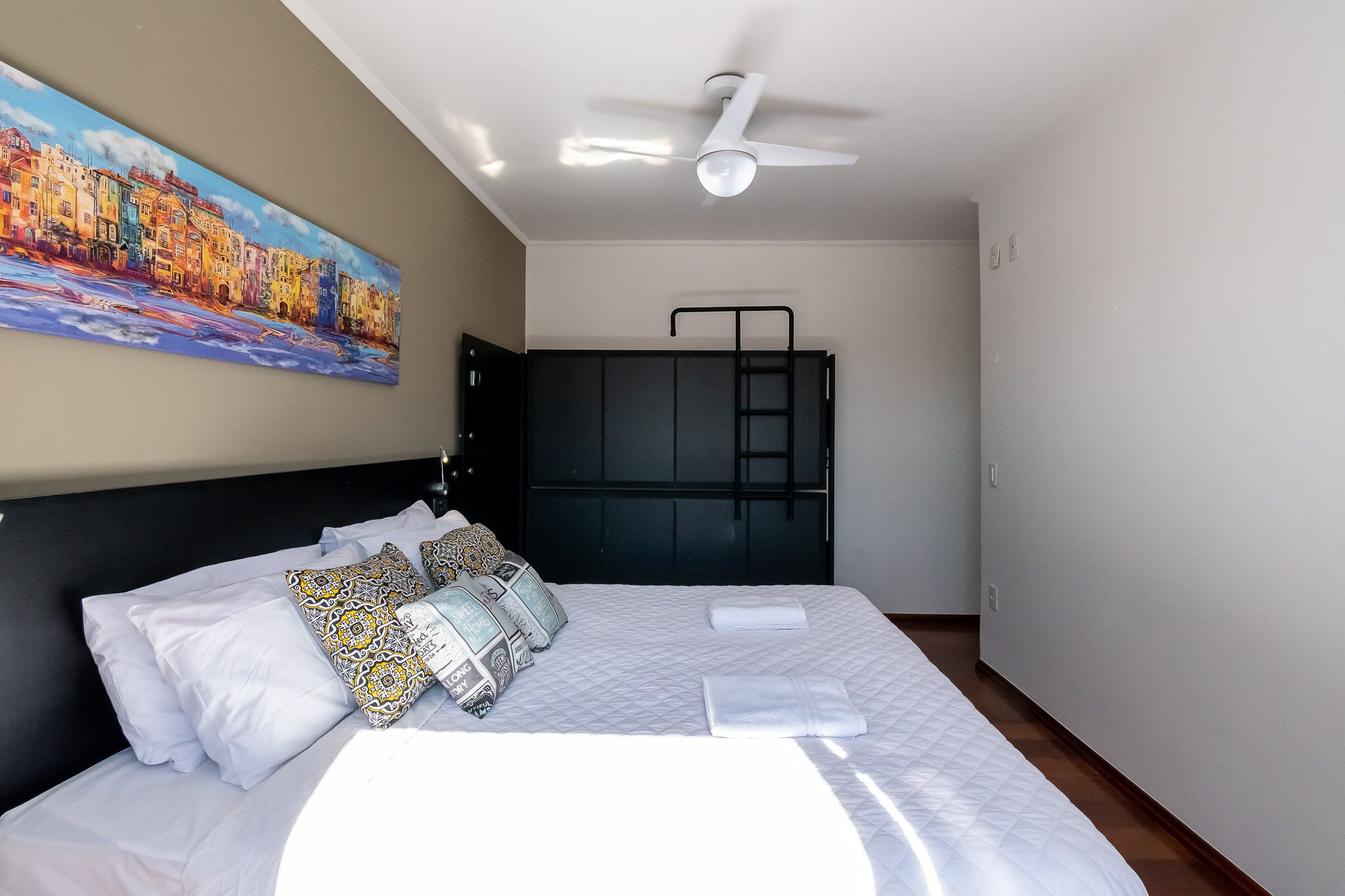 A neatly made bed with white linens and patterned pillows, a ceiling fan, and a painting of a colorful cityscape on the wall.