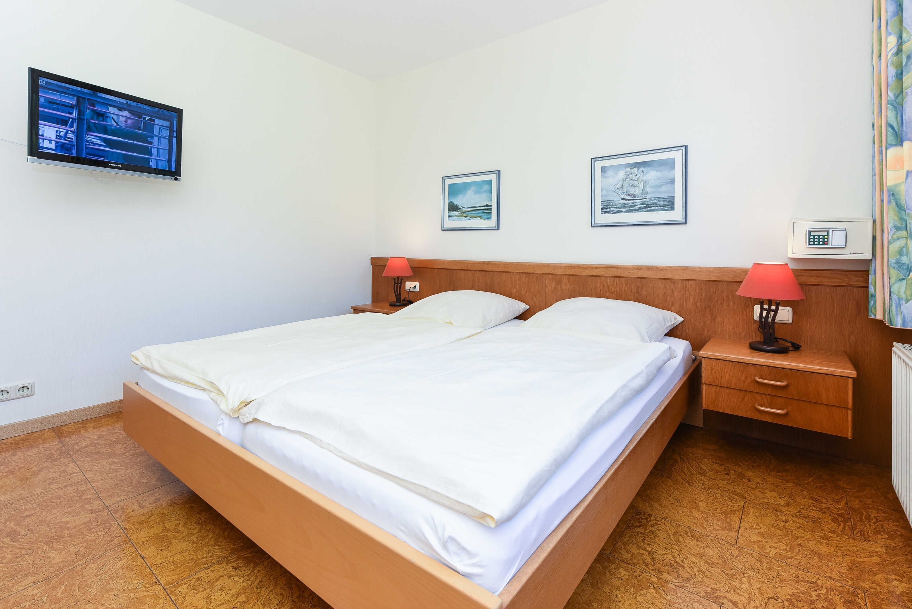 Une chambre avec un lit simple et un cadre en bois, deux tables de chevet avec des lampes, un téléviseur à écran plat fixé au mur et deux tableaux encadrés accrochés au mur.