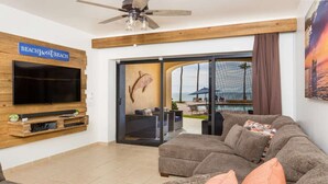 A living room with a flat-screen TV mounted on a wooden wall, a ceiling fan, and a view of an outdoor pool area.